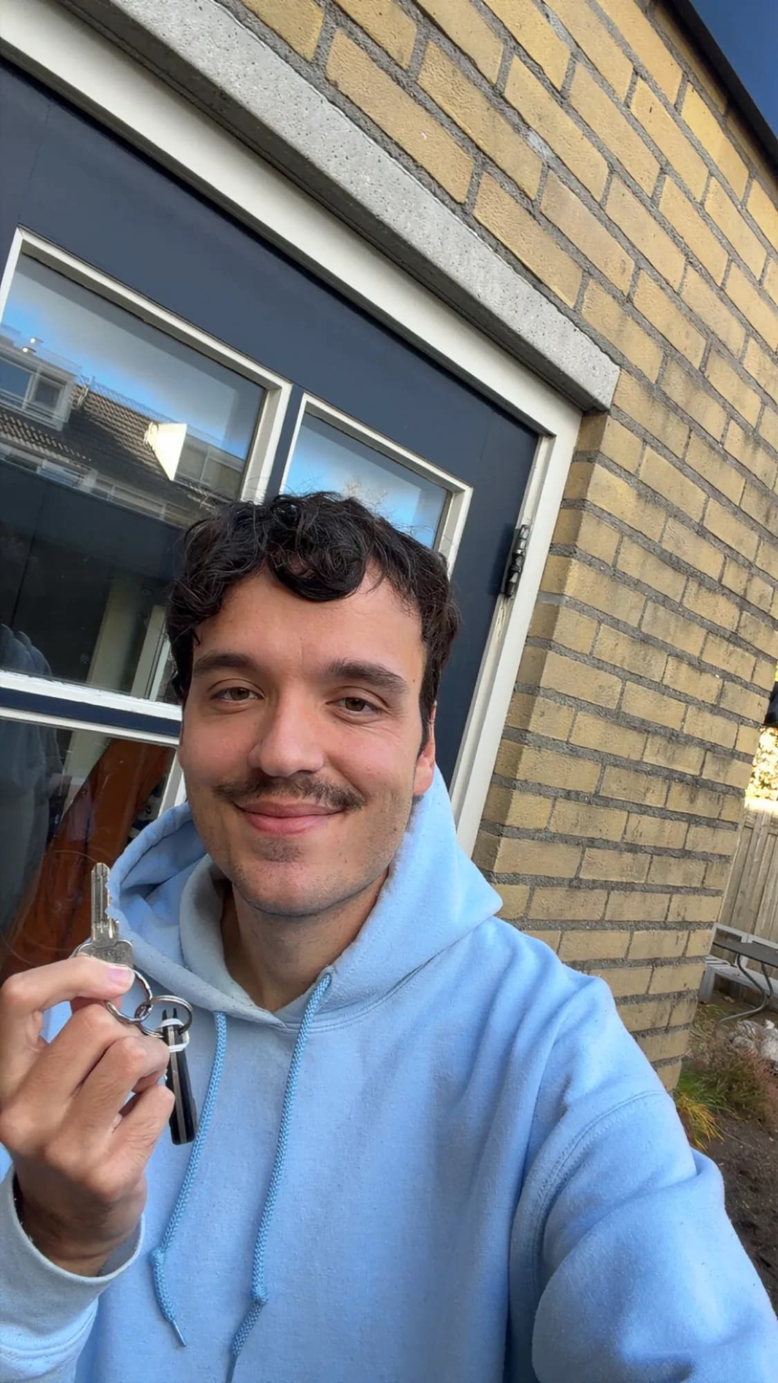 Lachende man in een lichtblauwe hoodie houdt een sleutelbos omhoog voor een voordeur